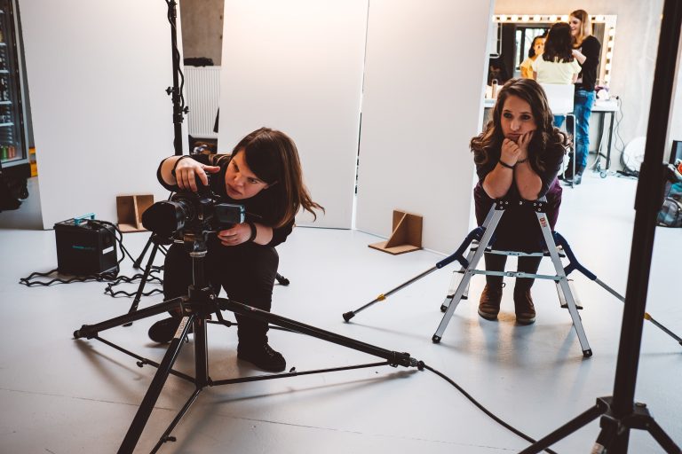 Fotografin mit Behinderung richtet ein Bild im Fotostudio ein. SIe steht hinter der Kamera und dem Stativ. Ihre Assistintin schaut ihr zu.