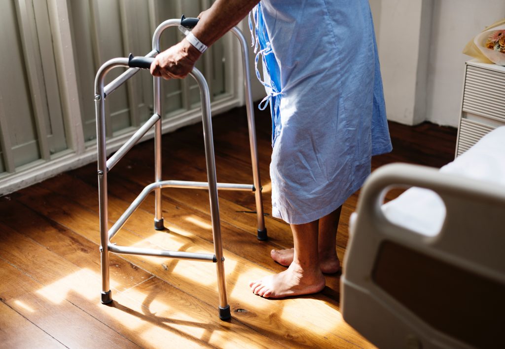 Ein älterer Mensch neben einem Krankenhausbett an einem Gehgestell sich aufstützend.