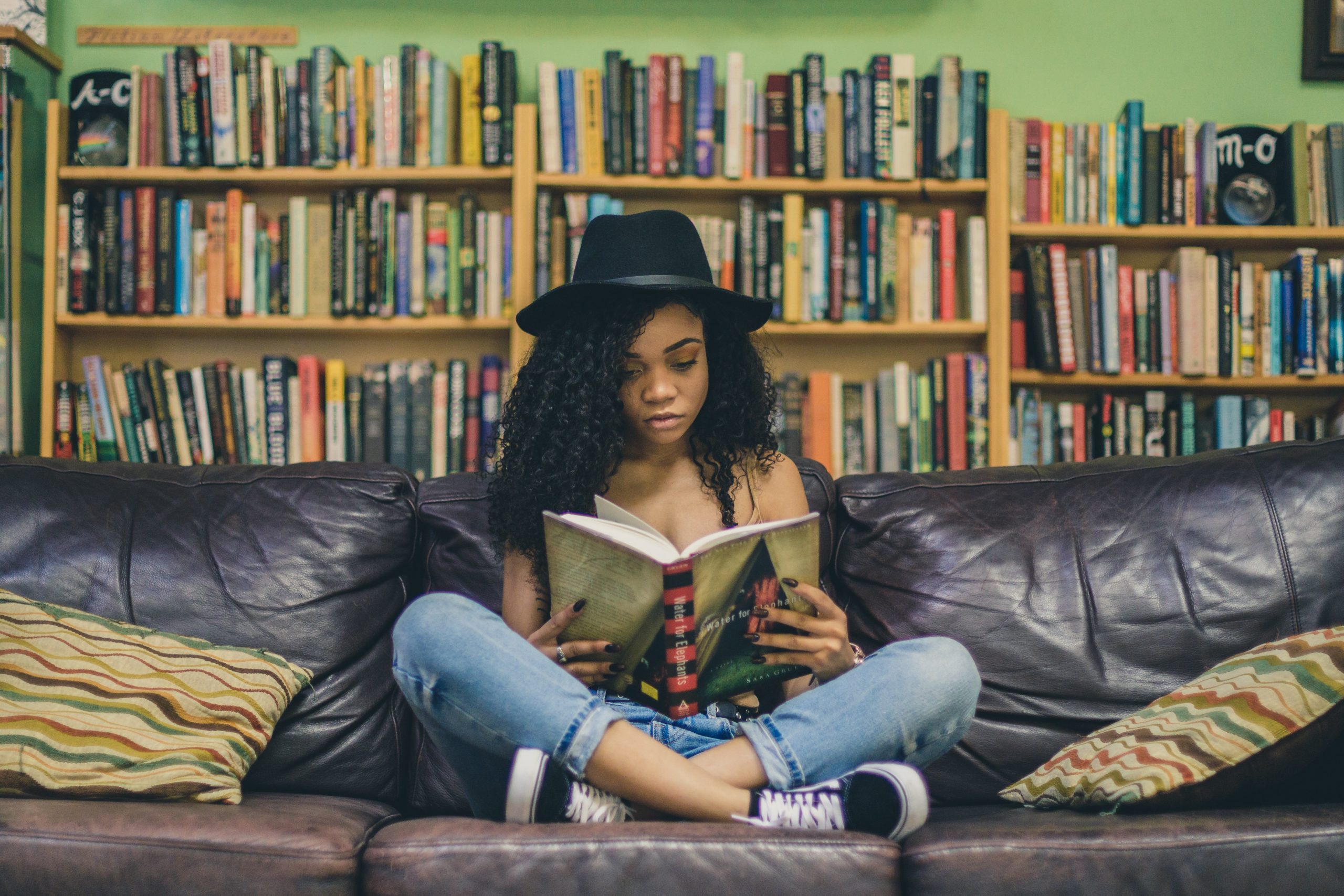 Eine Schwarze junge Frau, die im Schneidersitz auf einem Sofa vor einem riesen Bücherregal sitzt. Sie ist vertieft in ein Buch, was sie in ihren beiden Händen aufgeschlagen hat.