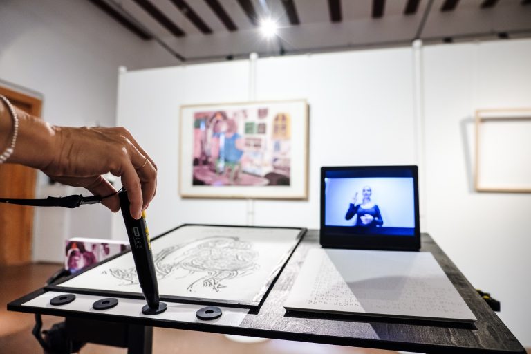 Es ist ein Ausstellungsraum, ein gerahmtes Bild hängt im Hintergrund an der Wand. Auf Tischhöhe steht ein Fernseher auf dem eine Person zu sehen ist, die Gebärdensprache spricht. Davor ist eine Tafel in Brailleschrift. Im Vordergrund ist eine weiße Hand mit Falten und Schmuck zu sehen, die einen Tiptoi in eins von vier dafür vorgesehene Anschlüsse hält.